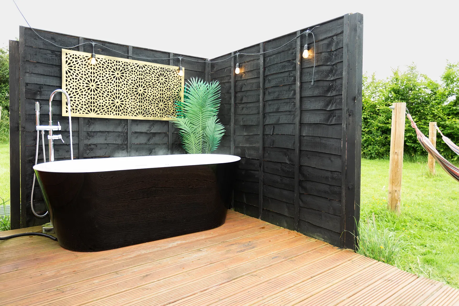 A black bathtub is sitting on a wooden deck next to a black fence.