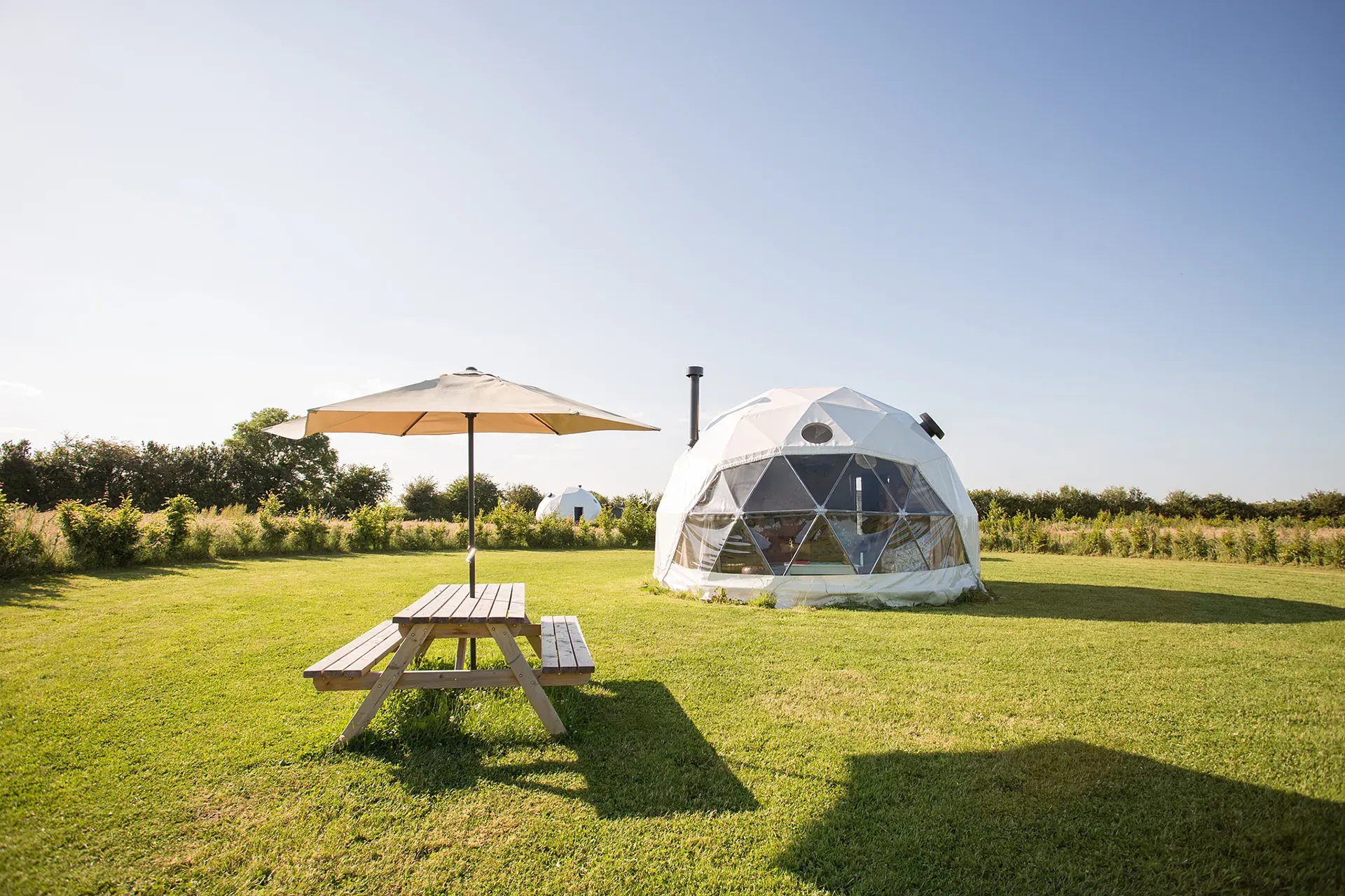 A dome tent is sitting in the middle of a grassy field next to a picnic table and umbrella.