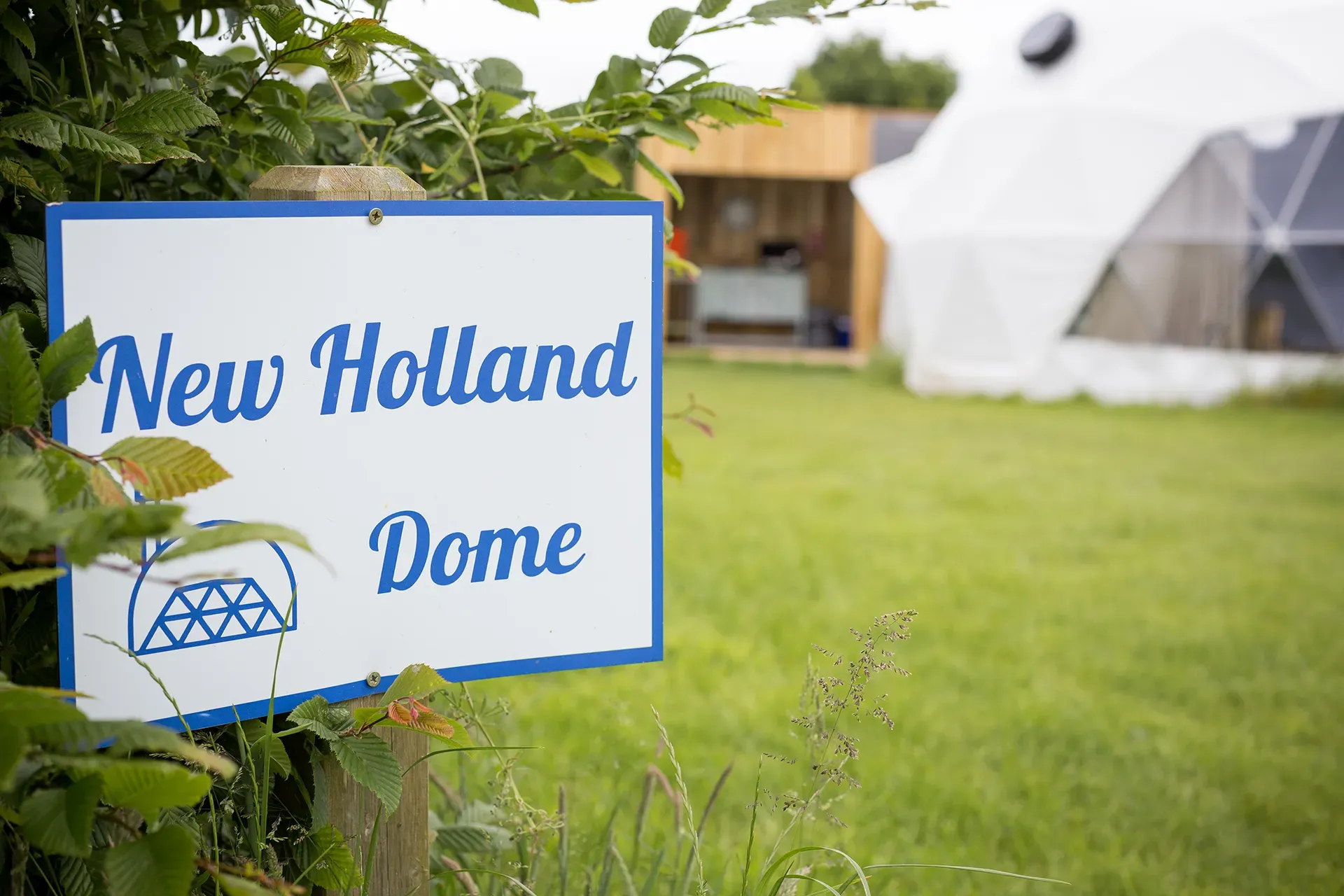 A sign for the new holland dome is sitting in the grass.