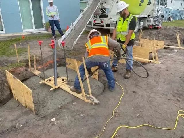 Construction workers pouring concrete into a formwork. Orange vests, white helmets, concrete truck, outdoor setting.