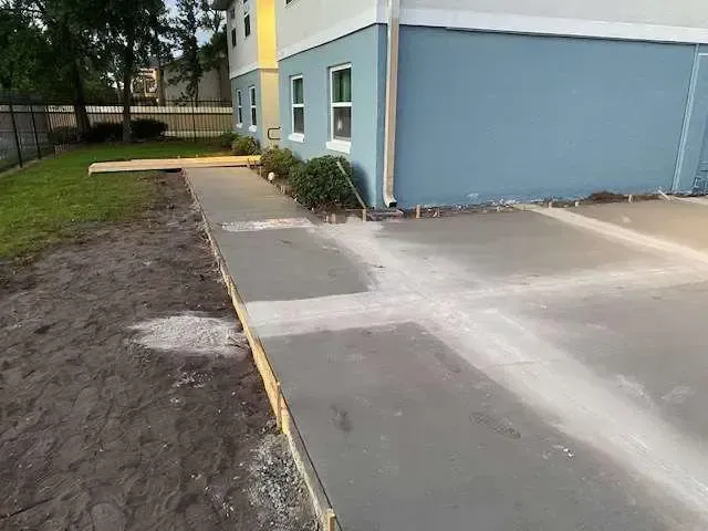 Fresh concrete sidewalk next to a light blue building with a wooden barrier. Grass and dirt surround.