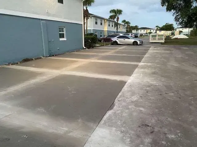 Newly poured concrete parking area at a multi-unit building with parked cars in the distance.