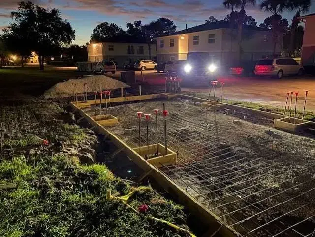 Construction site at dusk. Forms, rebar, and a backhoe in front of buildings.