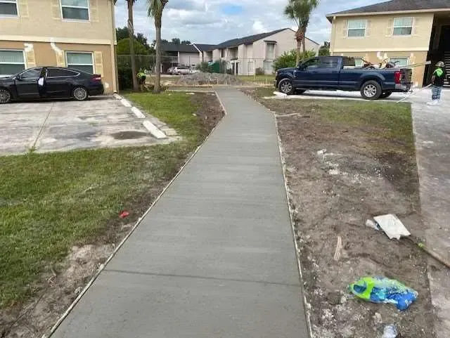 New concrete sidewalk in front of apartment buildings. Trucks and construction materials present.