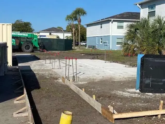 Construction site with concrete foundation, rebar, and wooden forms; buildings in background.