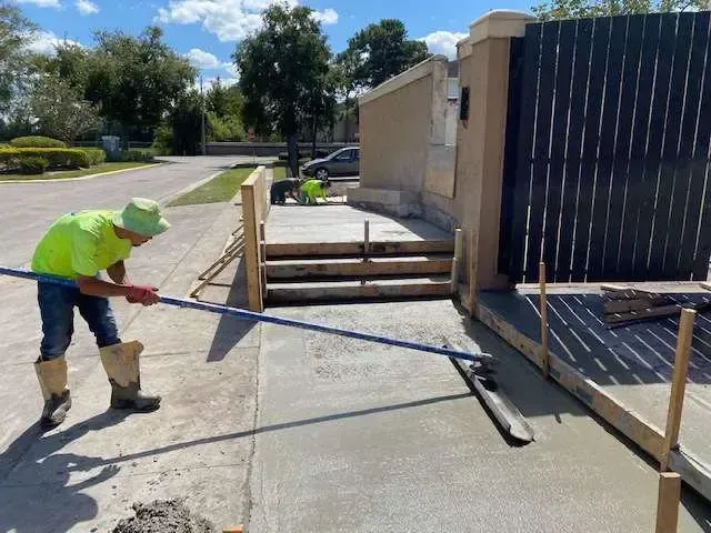 Construction worker smoothing wet concrete driveway with a long tool.