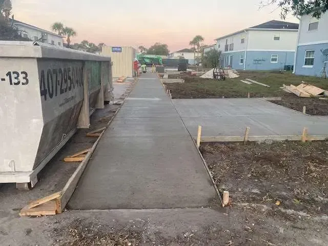 Newly poured concrete sidewalk and adjacent pad at a construction site; dumpsters and buildings in the background.