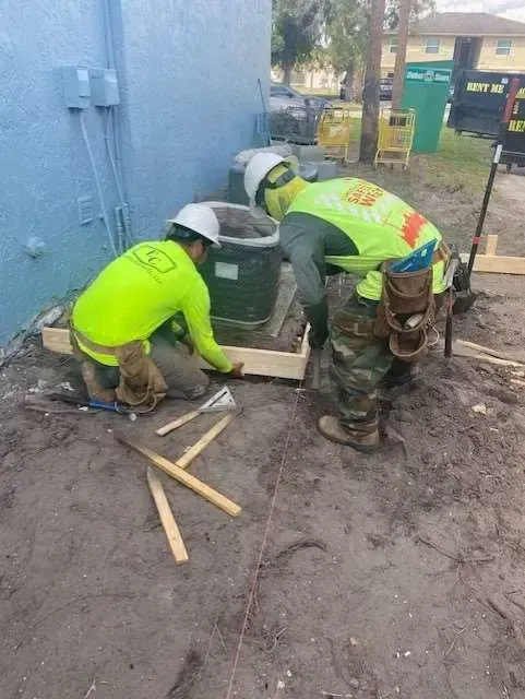 Two construction workers installing a wooden frame around an AC unit outdoors.