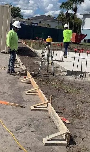 Construction workers setting concrete forms outdoors with a laser level, wearing hard hats and safety vests.