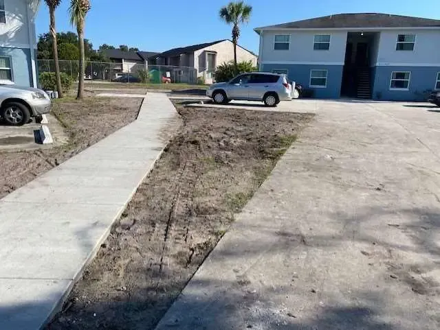 Concrete sidewalks and driveway border a dirt patch in front of a two-story apartment building.