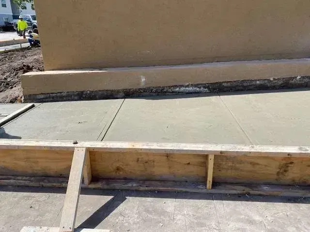 Construction site with a concrete sidewalk and wooden formwork, with a tan wall in the background.