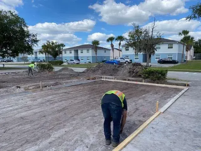 Construction workers building a foundation for a structure outdoors. They wear safety vests. Apartment buildings and blue sky in background.