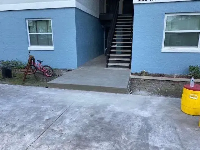 Exterior view of an apartment building with a concrete ramp leading to stairs. Blue walls, bike, and yellow barrel.