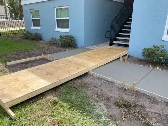 Wooden ramp providing accessibility to an apartment entrance; new concrete path next to ramp, blue building.
