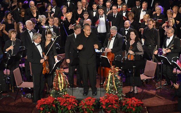 Orchestra and choir performing on stage, with poinsettias, conducting.