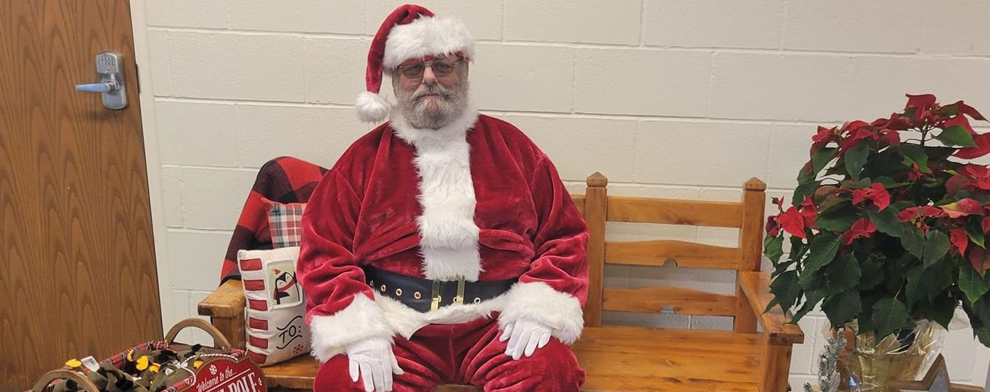 Santa Claus sits on a wooden bench surrounded by gifts and poinsettias.