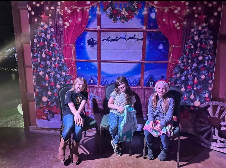 Three young girls sitting, posing in front of a Christmas backdrop with a window, trees, and Santa's sleigh.