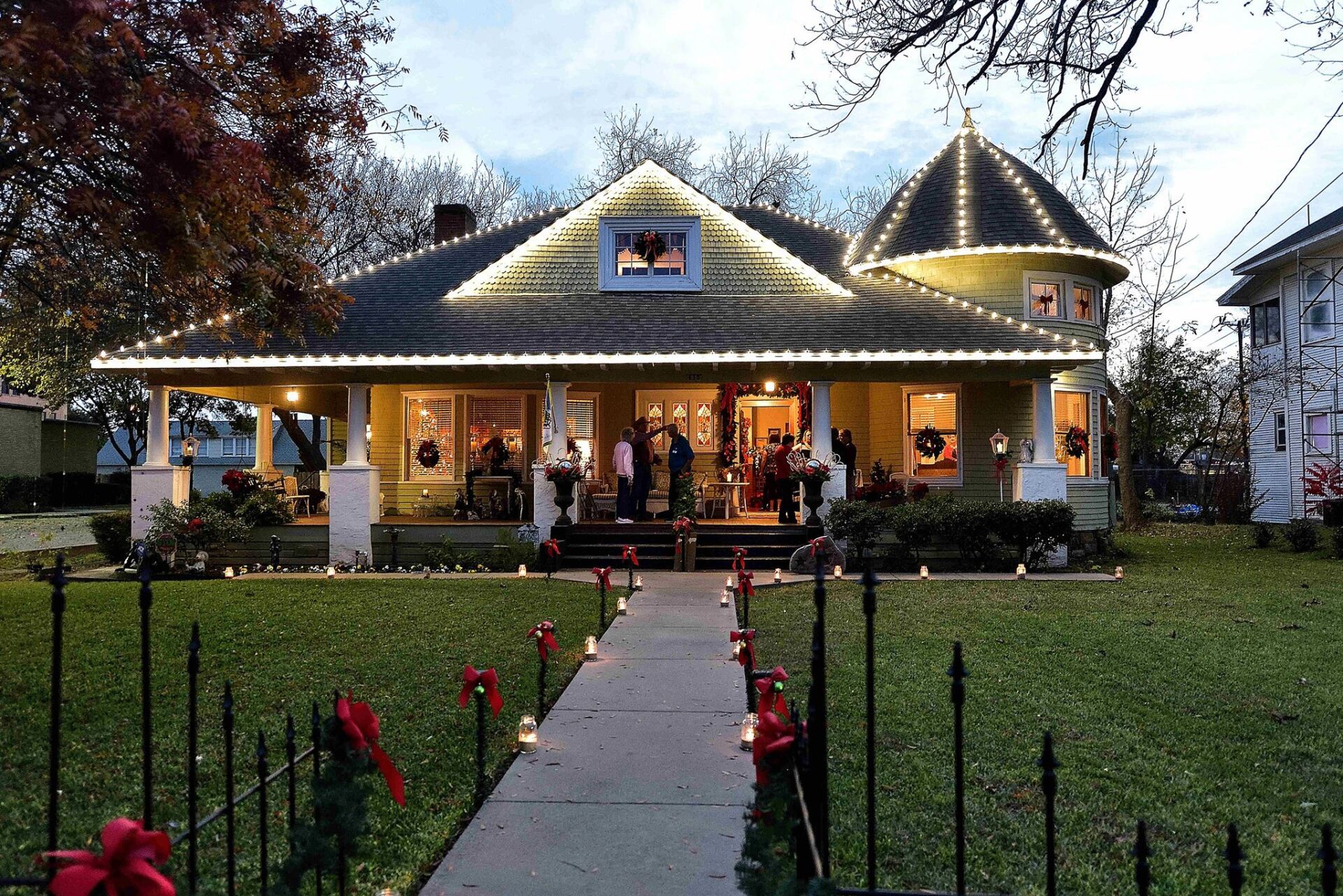 Festive house decorated with lights and red bows on a sidewalk, people gathered on the porch, evening scene.