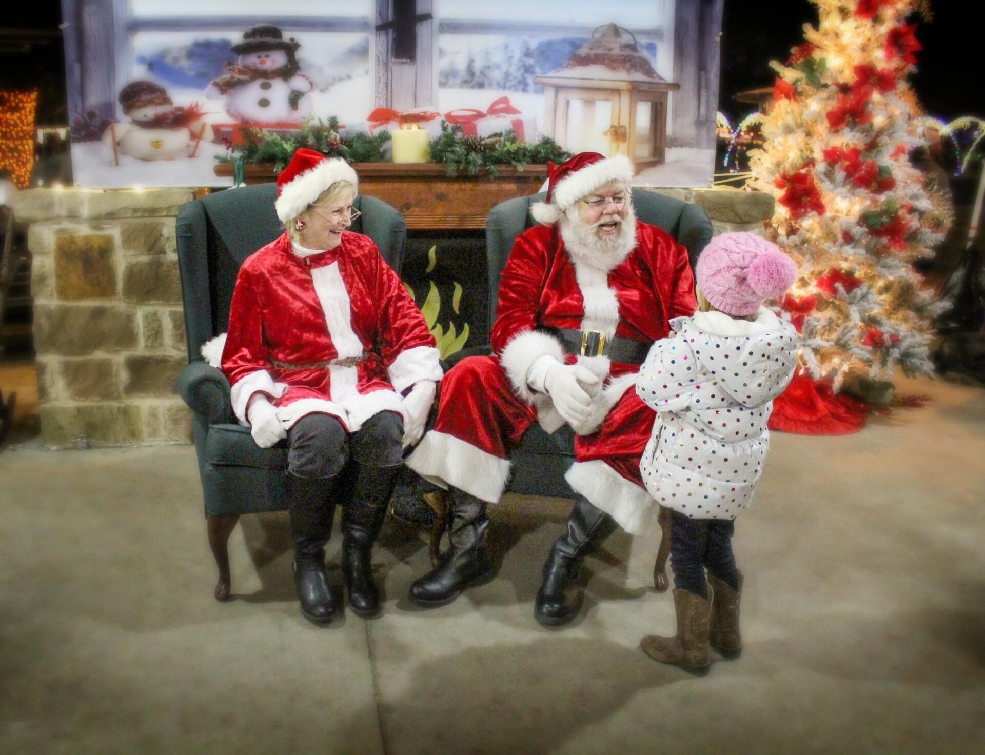 Santa and Mrs. Claus sit near a fireplace as a child in a pink hat talks to them.