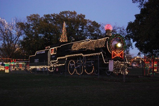 A black train decorated with Christmas lights sits on a grassy lawn at dusk.