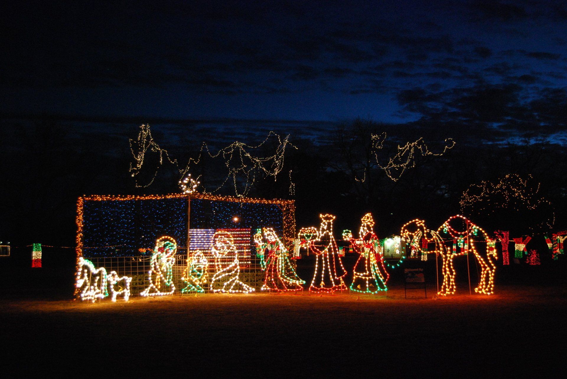 Nativity scene with Christmas lights, figures of Mary, Joseph, wise men, camel, and baby Jesus.