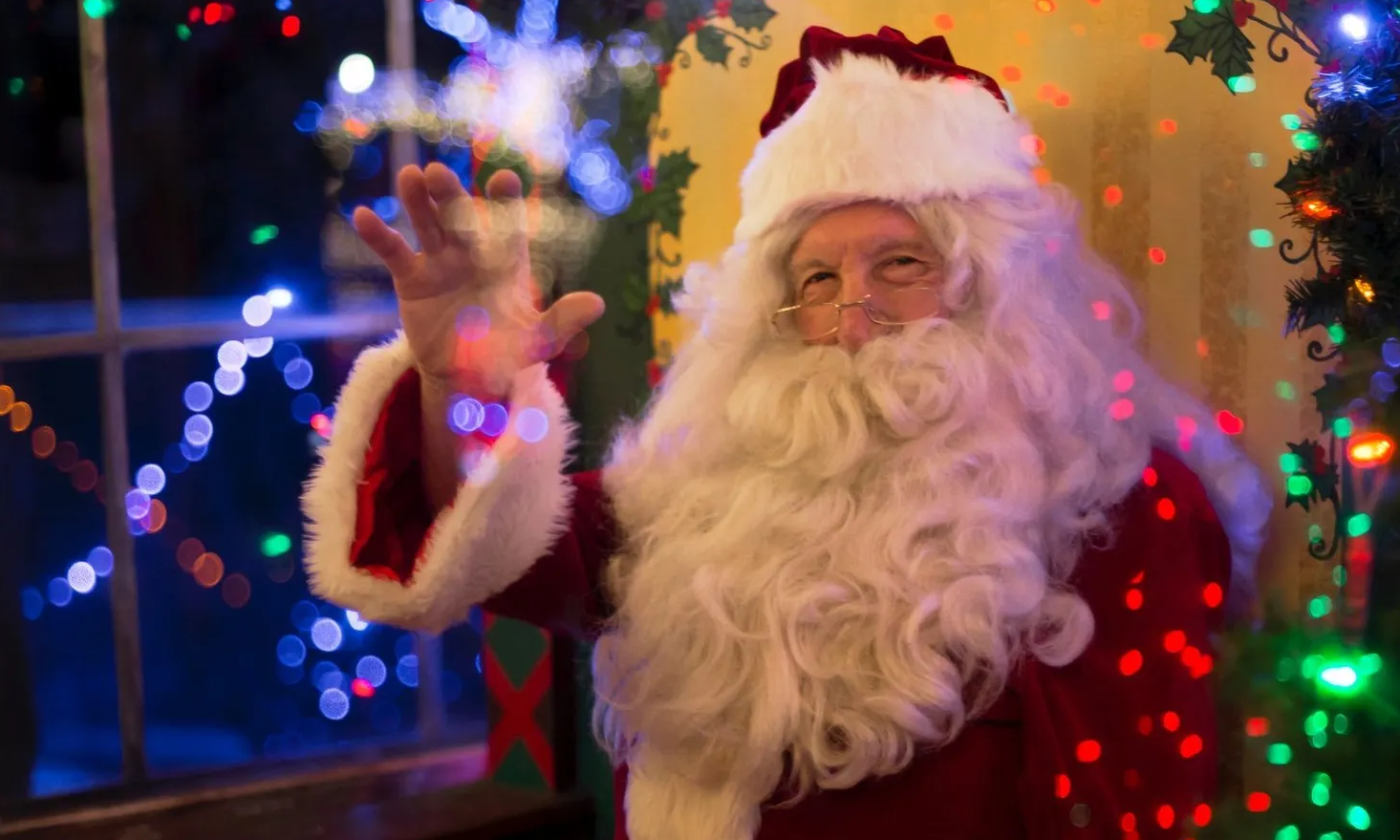 Santa Claus, in red suit, waving and smiling, sits by decorated Christmas tree and window.