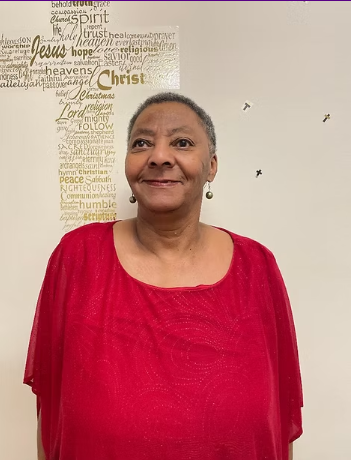 Woman in red top smiles in front of a wall with a cross-shaped religious text and small bird silhouettes.