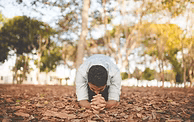 Man kneeling on fallen leaves, hands clasped in prayer, outdoors in a park setting.
