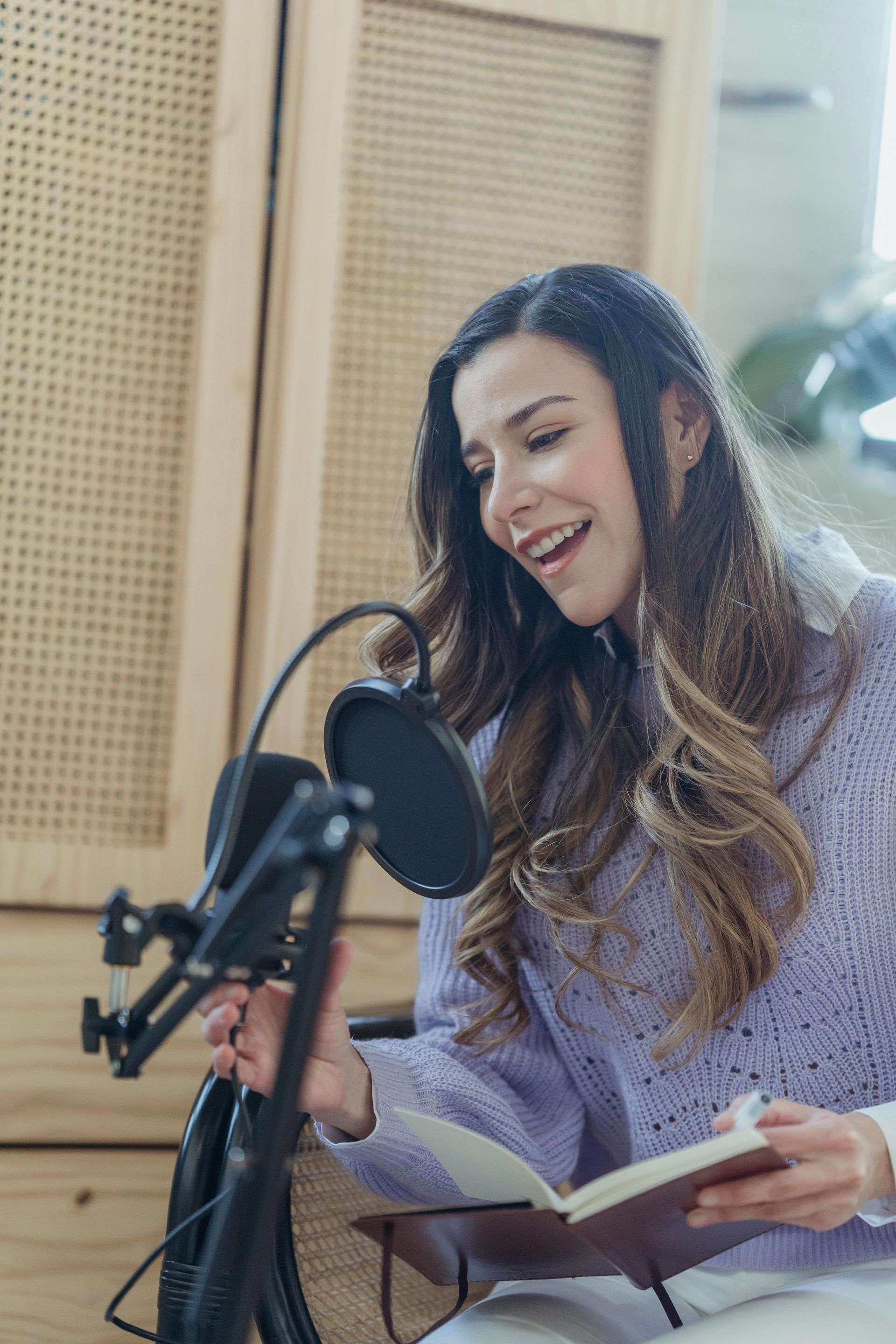 Woman speaking into a microphone, holding a notebook. Wearing a lavender sweater, in a studio setting.