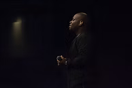 Man in dark clothing holding a microphone, standing on a stage with a spotlight.