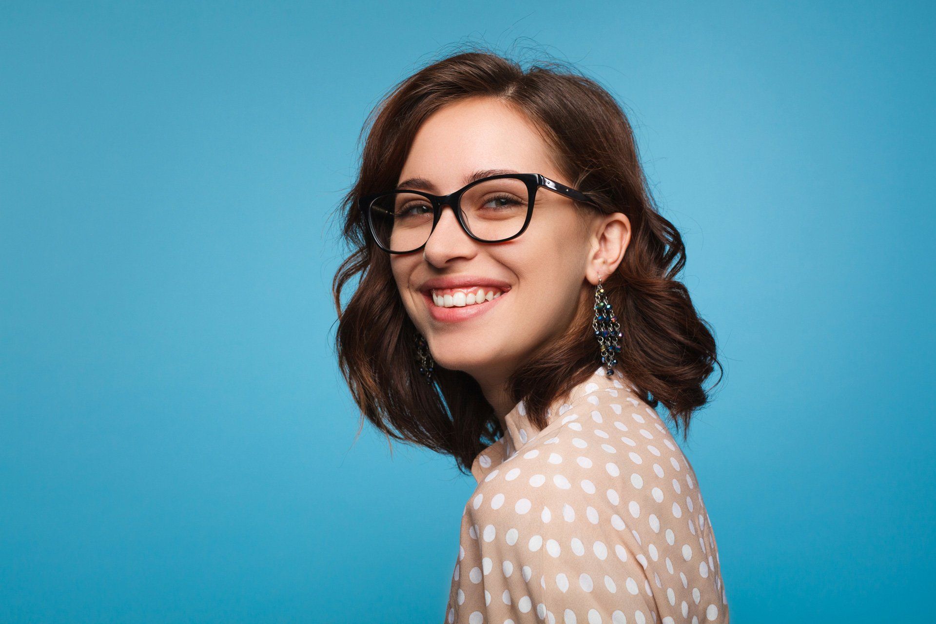 Woman with glasses smiles broadly, looking over her shoulder against a blue backdrop.