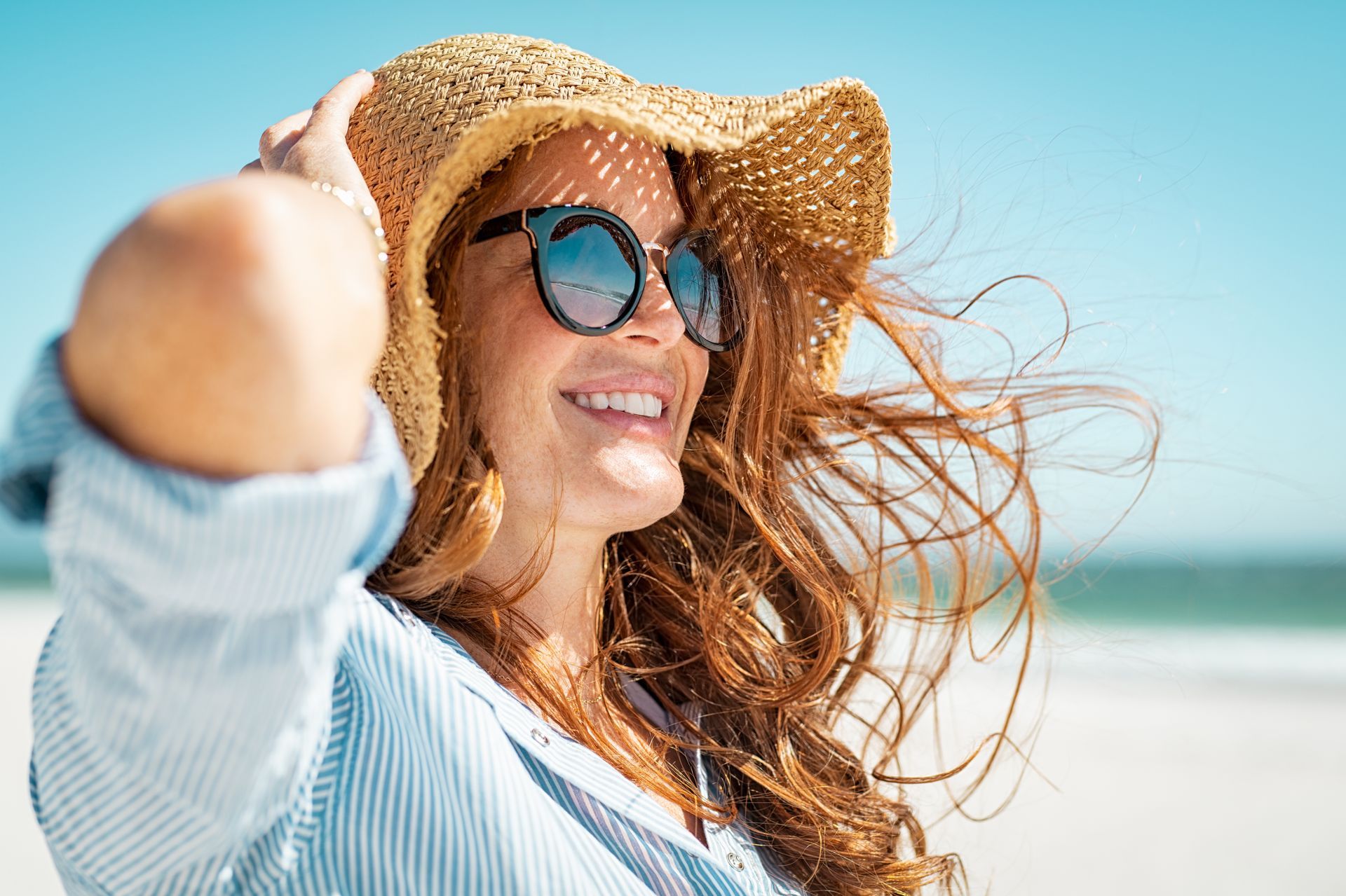 Person holding a sunhat on a windy beach with ocean and bright sky in the background.