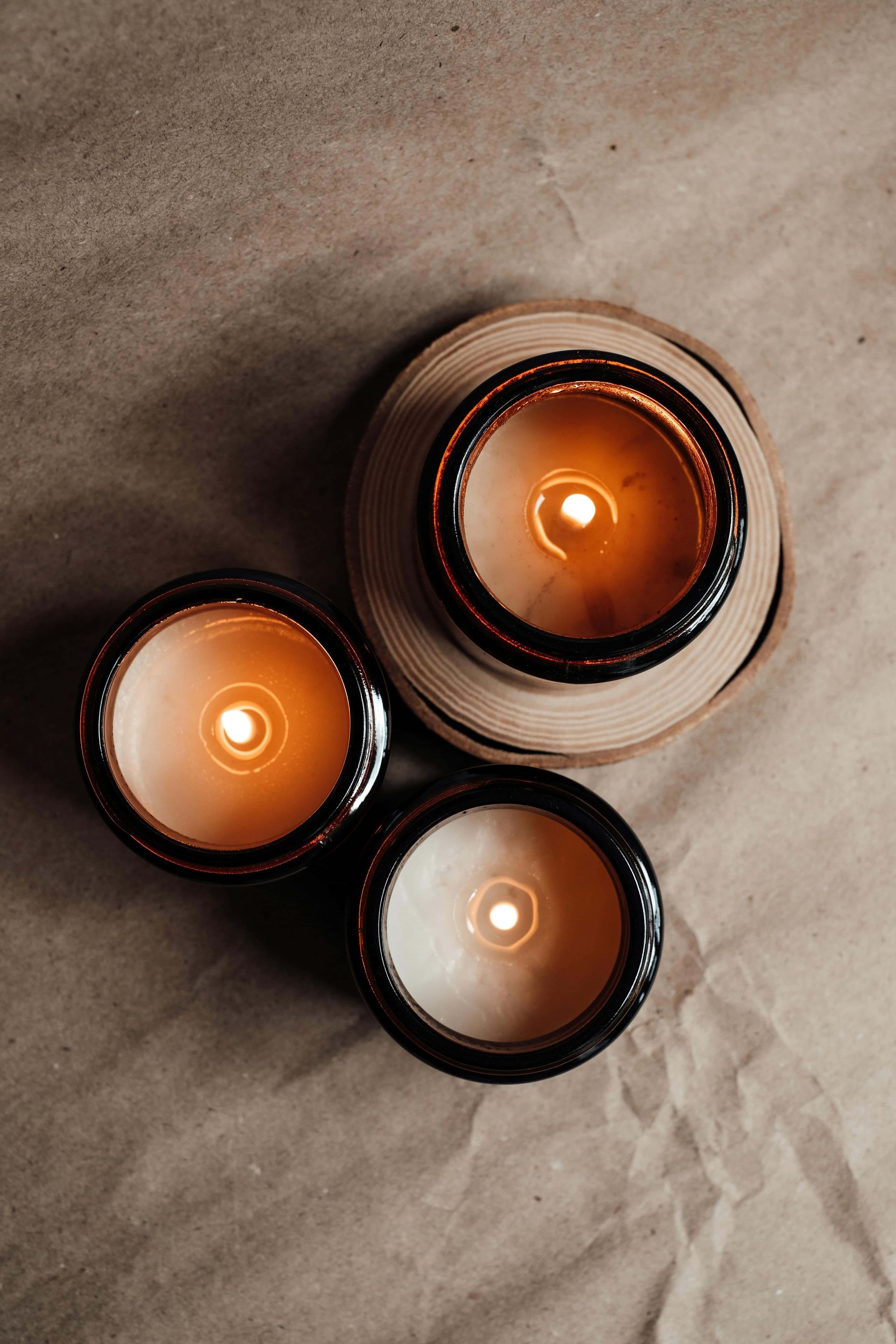Three lit candles in dark glass jars on a textured brown surface. The candles glow with warm light.