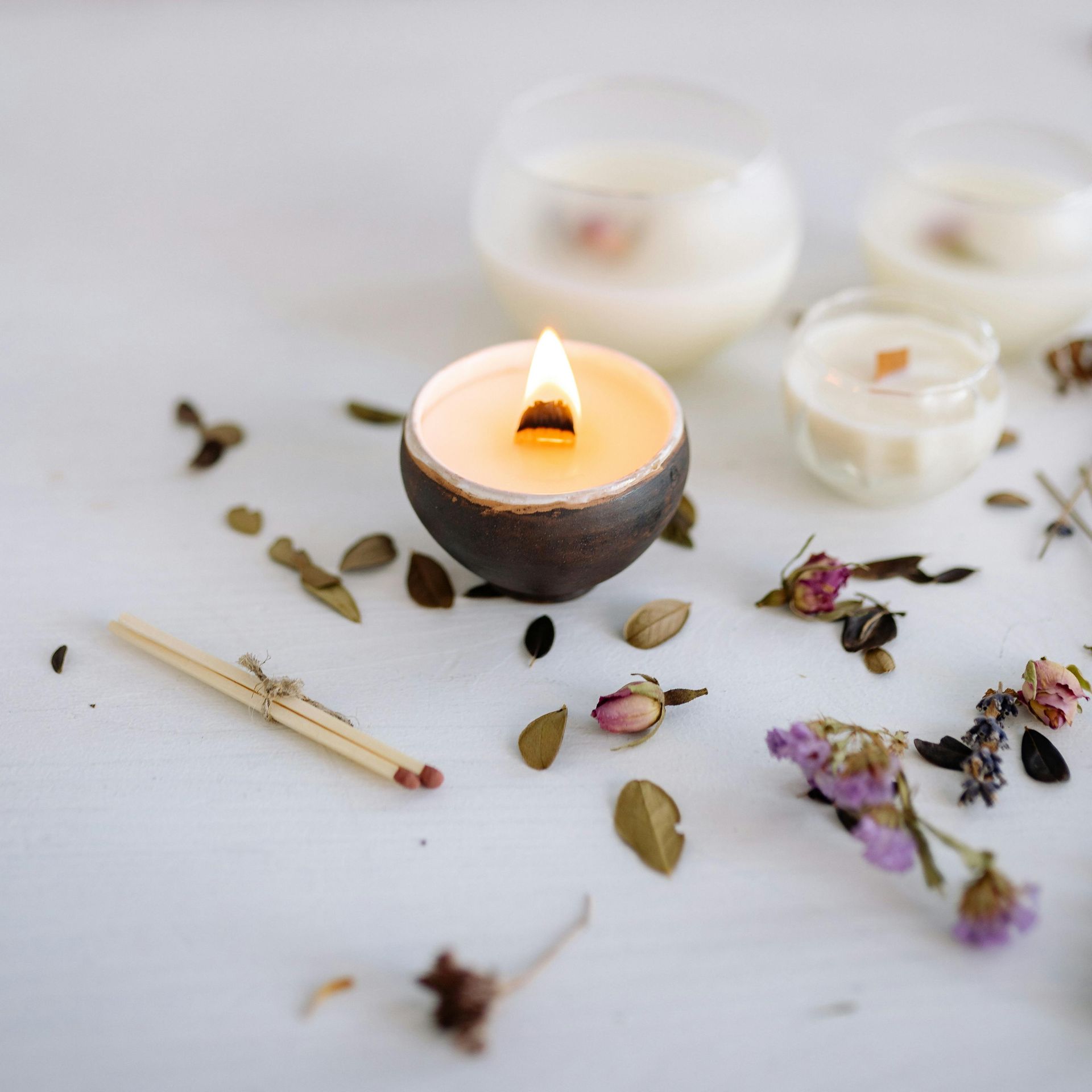 Lit candle in a ceramic bowl surrounded by dried flowers, leaves, and matches on a white surface.