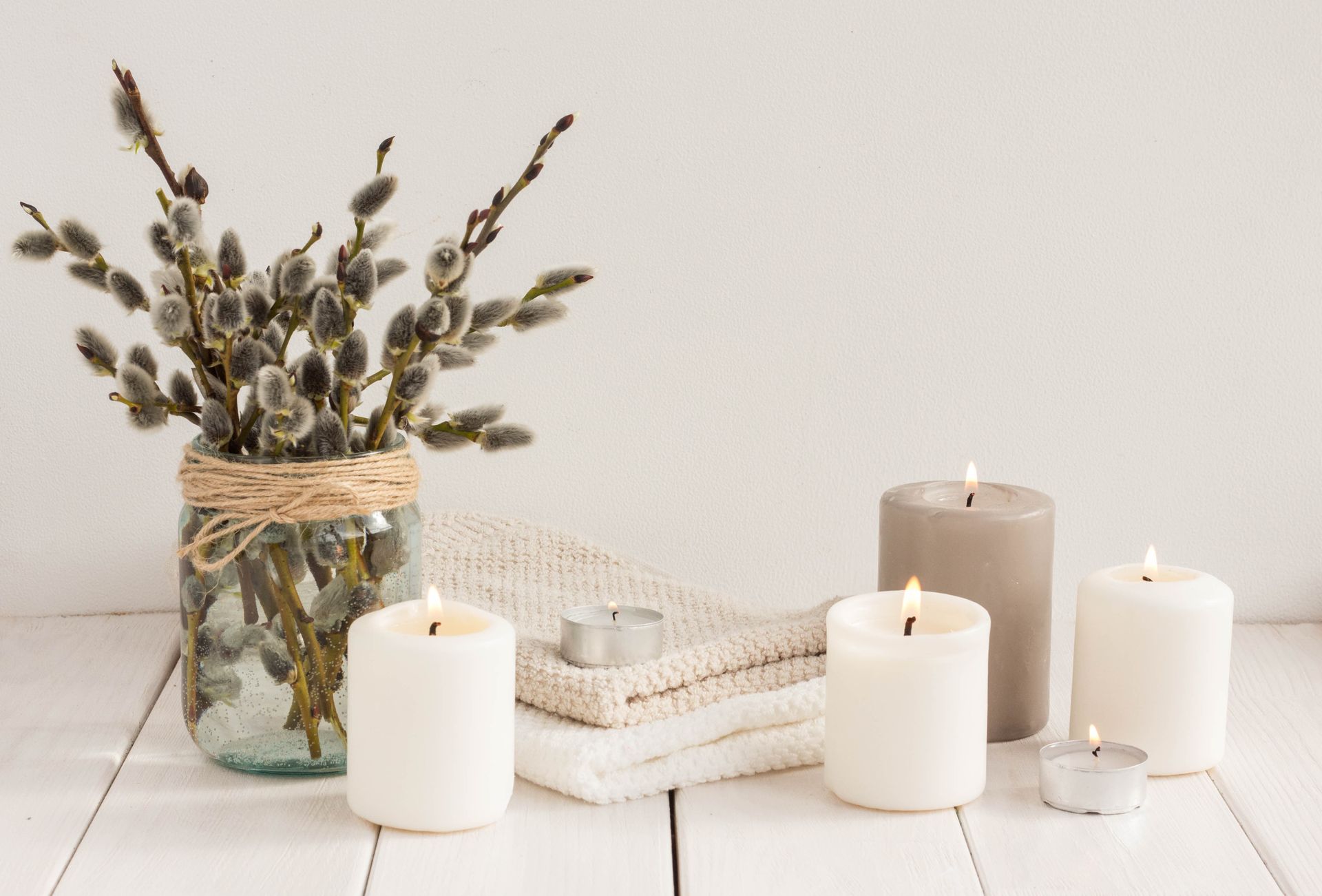 A vase of pussy willows with lit candles and a folded towel on a white wooden surface.