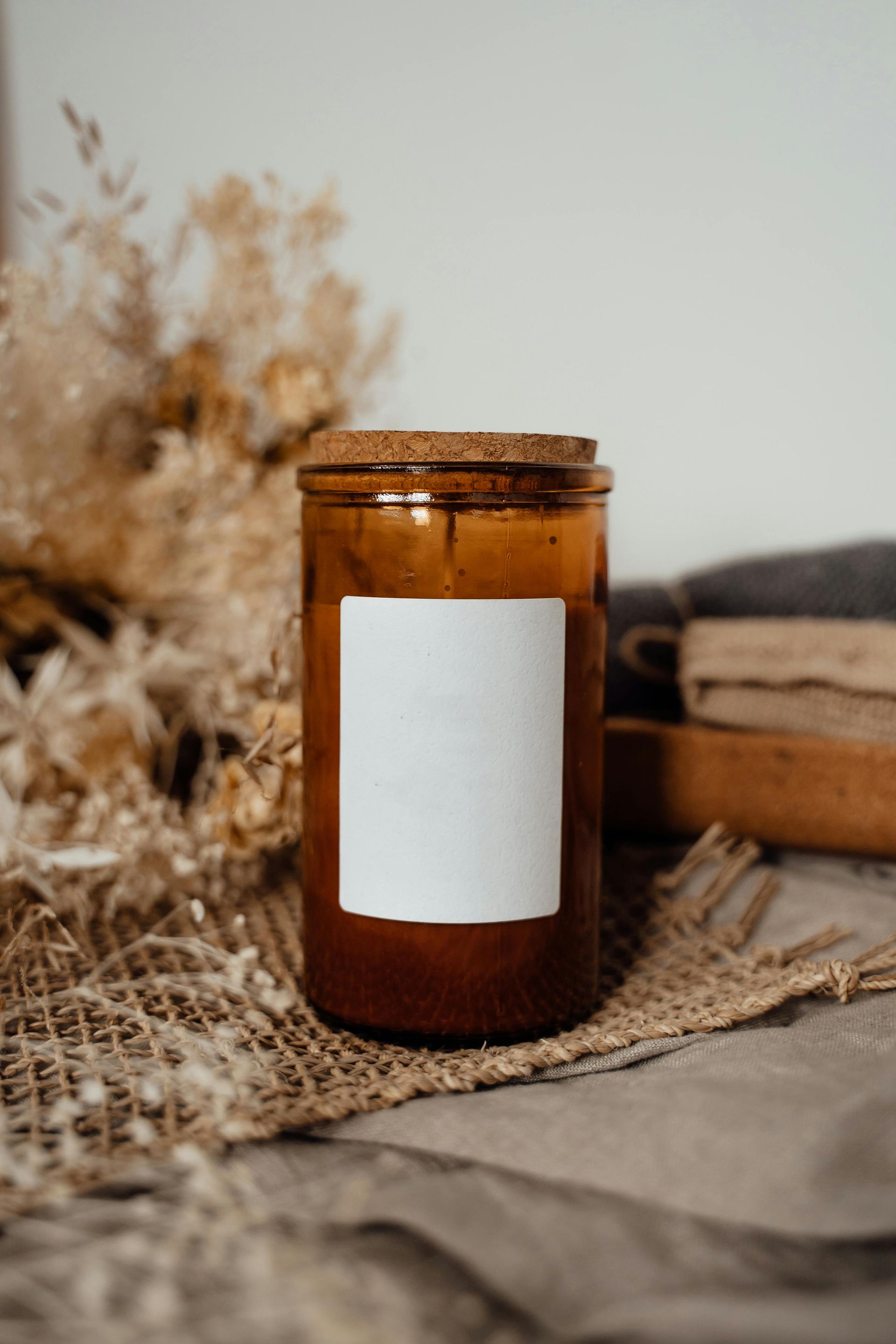 Amber glass candle jar with blank label, burlap, and dried flowers in soft light.