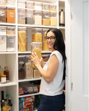 Woman in a pantry holding a jar of pasta beside neatly labeled storage containers.