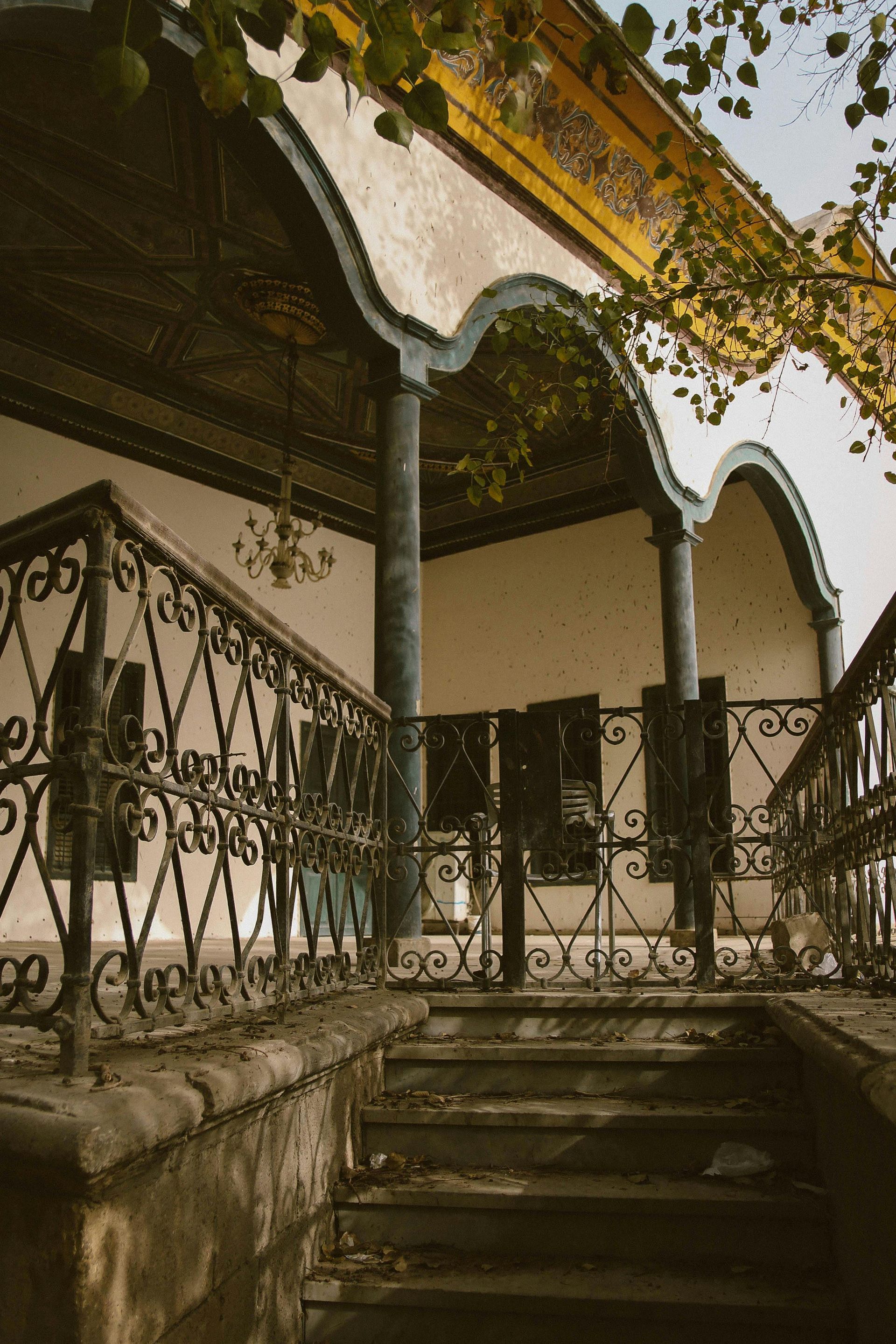 Stone steps lead to a porch with ornate metal railing and dark pillars. Overhead, a curved roof.