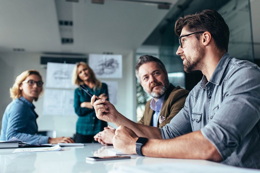 People in a meeting, discussing ideas. Man speaking, others listening at a table in an office.