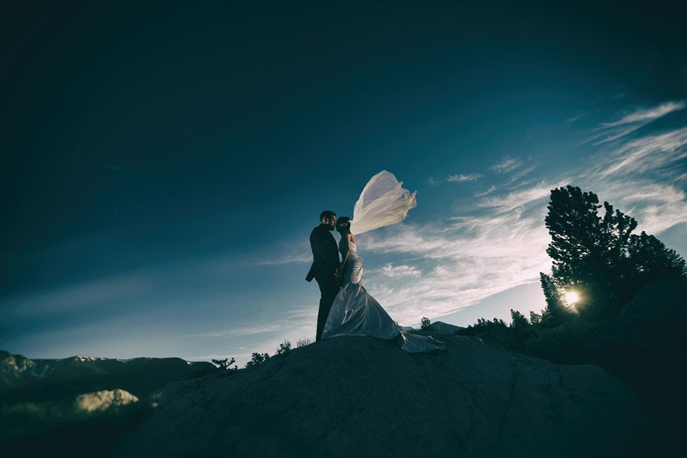 Bride and groom embrace on a mound; veil blowing in the wind against a blue sky with sun.