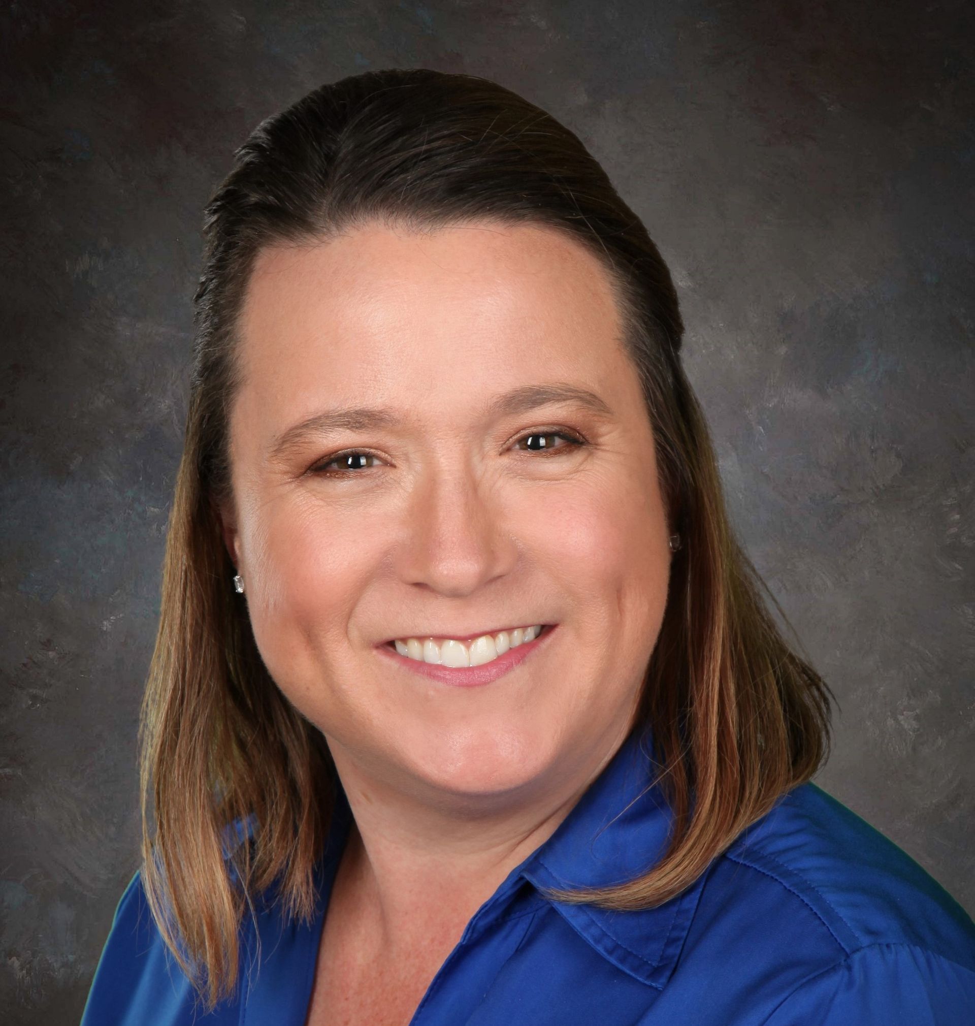 Woman smiling, wearing a blue shirt. Brown hair pulled back. Dark blurred background.