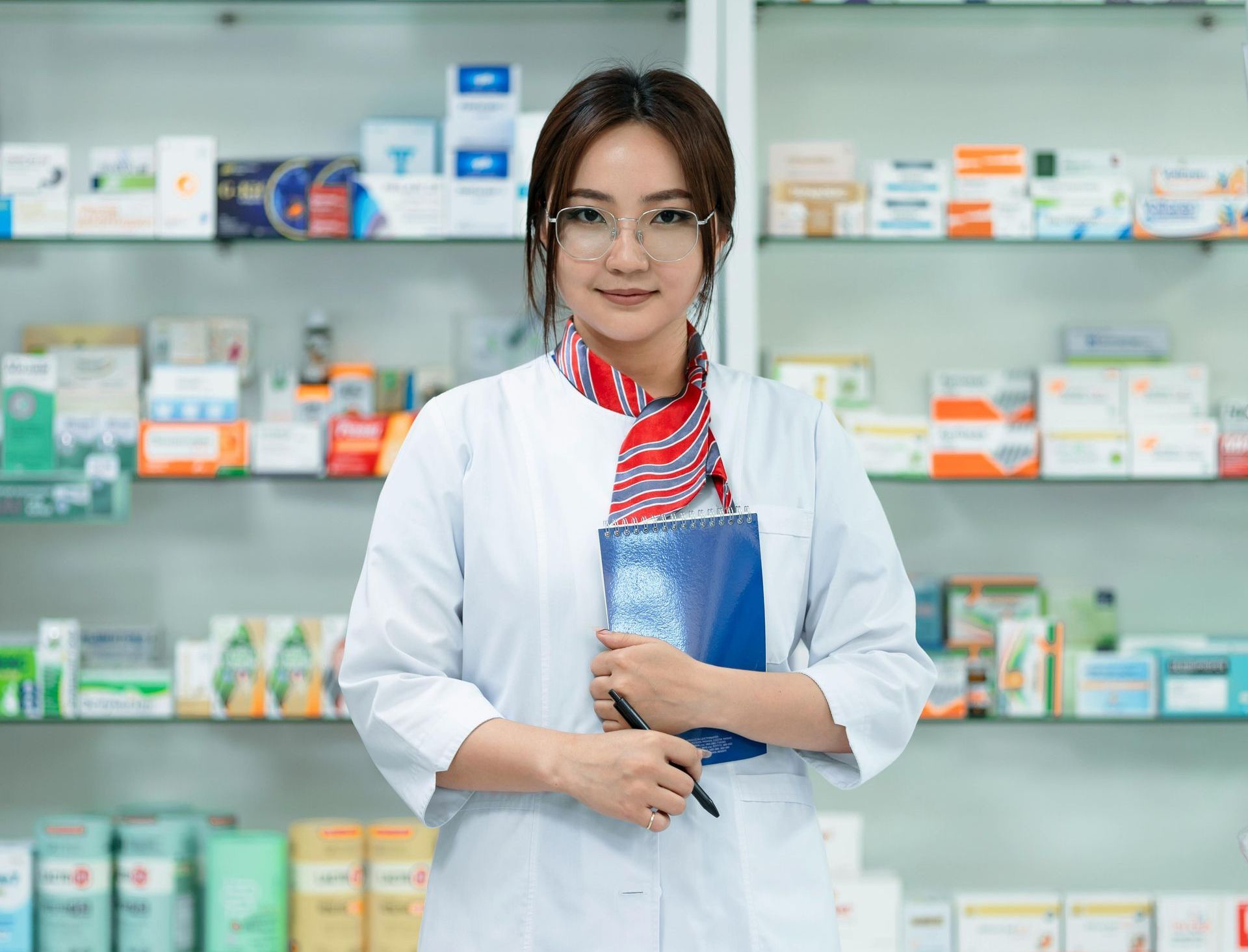 Pharmacist in white coat holding a blue notebook, standing in front of shelves of medicine.