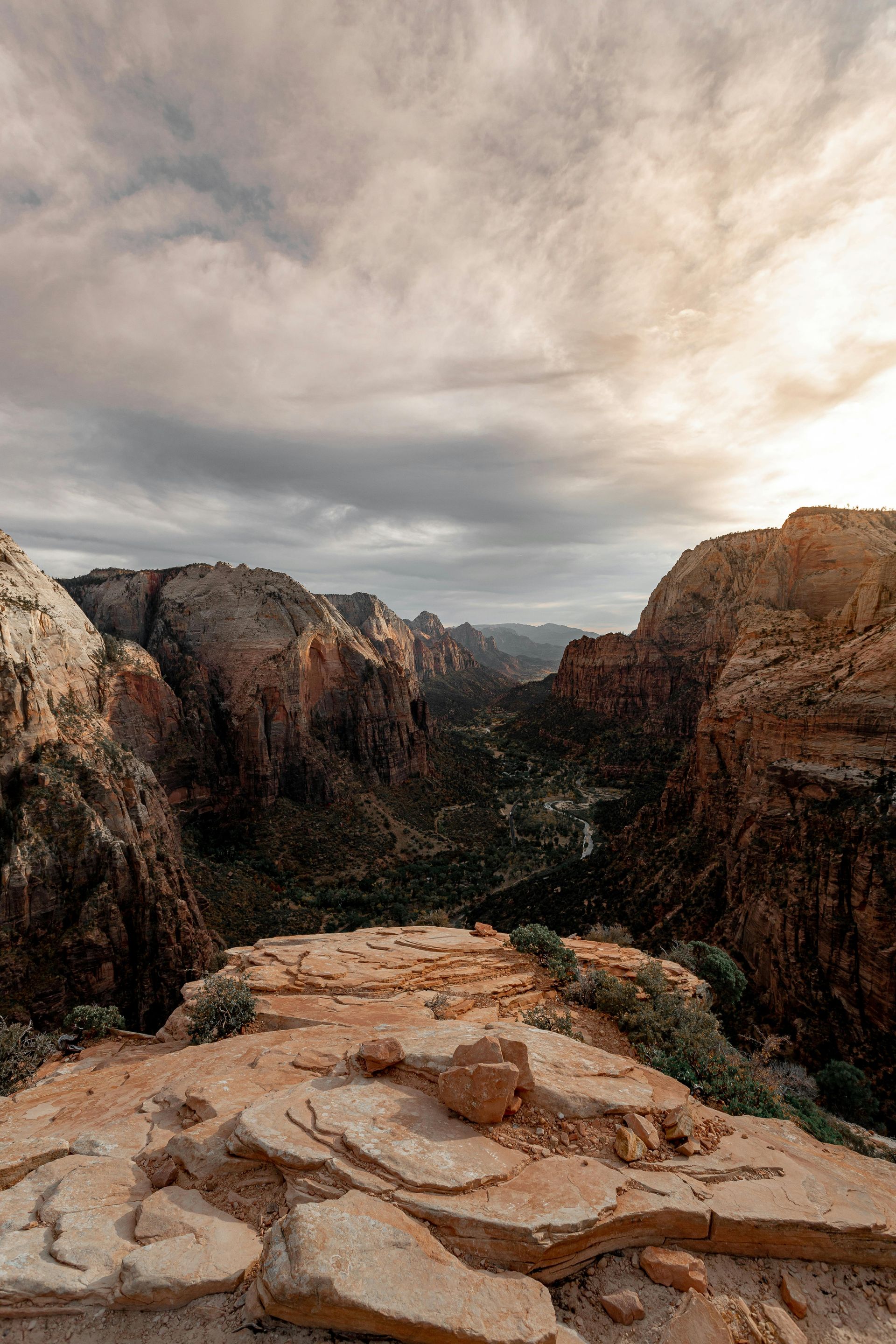 Canyon view: Rocky foreground overlooking a deep valley with reddish-brown cliffs and a cloudy sky.