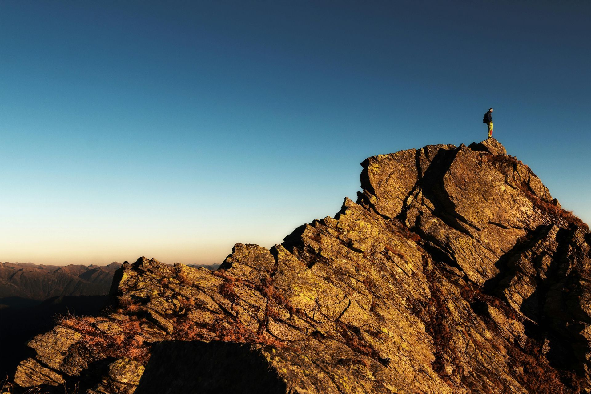 Person stands atop a rocky mountain peak against a clear blue sky.
