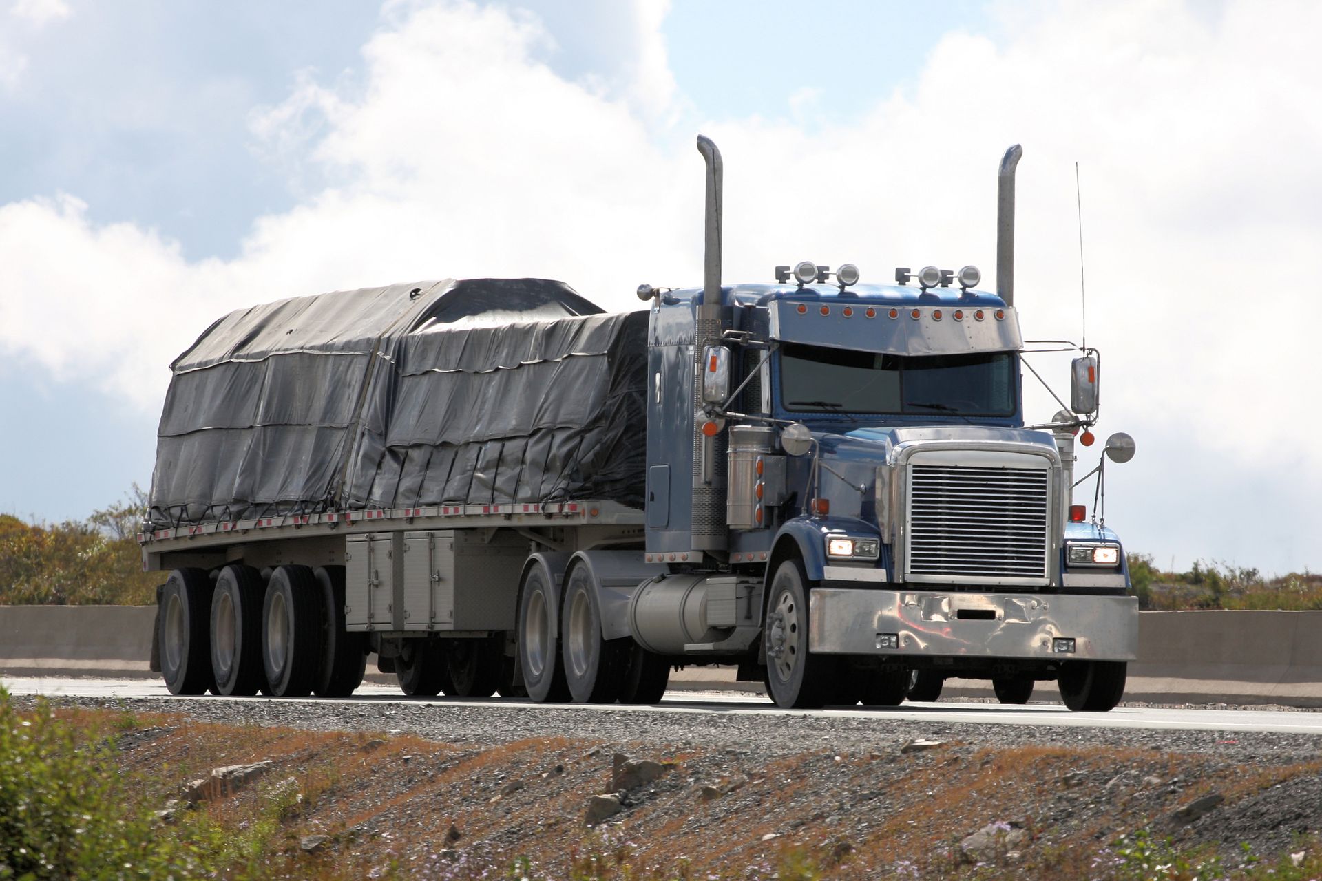 Blue semi-truck hauling a covered flatbed trailer on a highway.