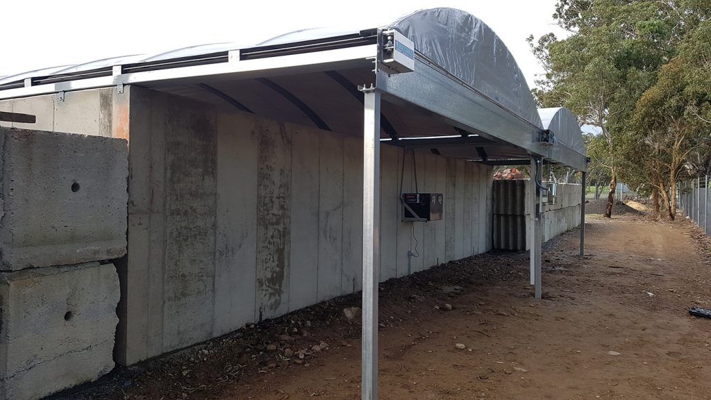 Greenhouse with metal frame and concrete walls; dirt path beside it.