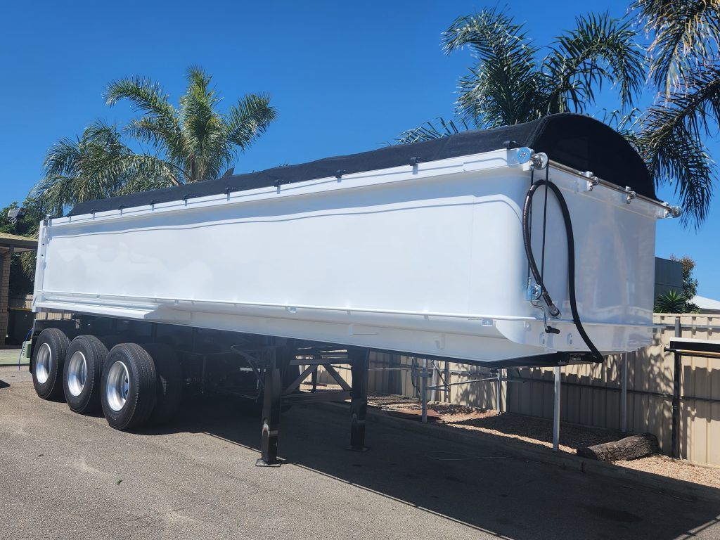 White semi-trailer with black tarp, parked outdoors under a blue sky.