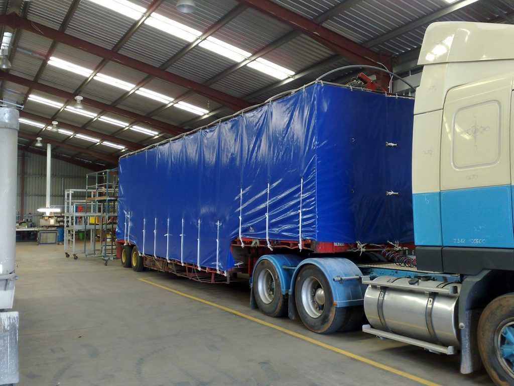 Blue tarp-covered semi-trailer inside a warehouse, connected to a truck with blue and white paint.