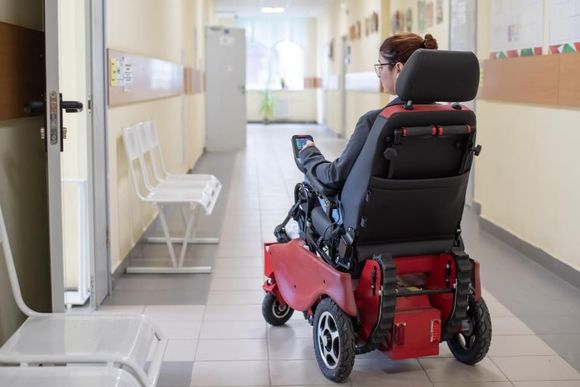 Woman in motorized wheelchair in a hallway, approaching a doorway. Red and black wheelchair. White seats on the left.