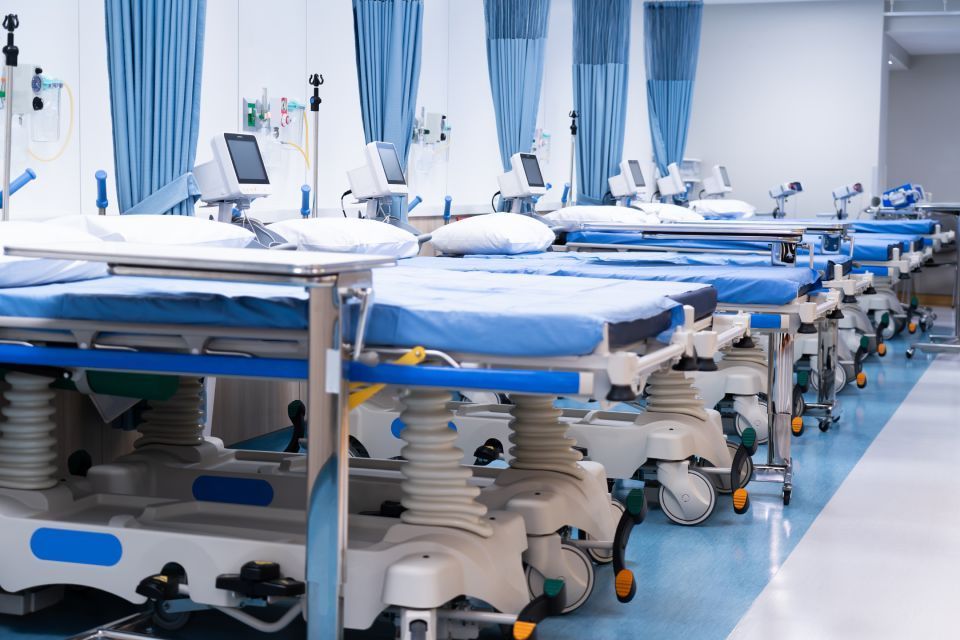 Empty hospital beds in a brightly lit room with blue curtains and medical equipment.